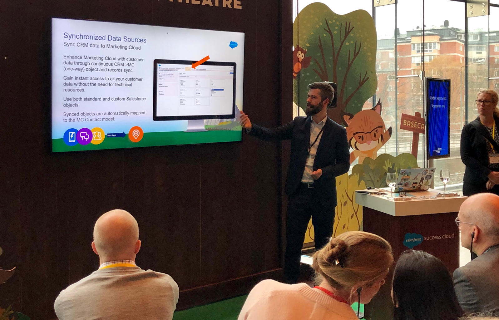 Presenter in a dark suit explains a slide titled 'Synchronized Data Sources' with a large monitor showing CRM data, audience watching in a conference room.