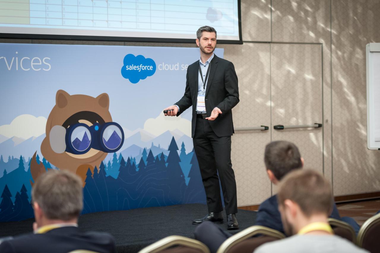 Male speaker in a suit presenting on stage at a conference, with a Salesforce banner and bear mascot backdrop behind him.