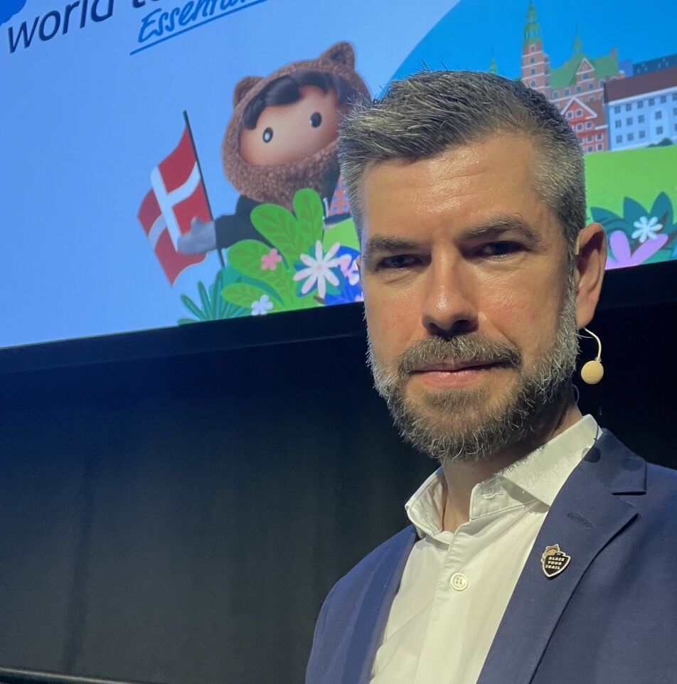 Male speaker on stage wearing a navy blazer and white shirt with a headset mic, backdrop shows a colorful screen with a mascot and flags.