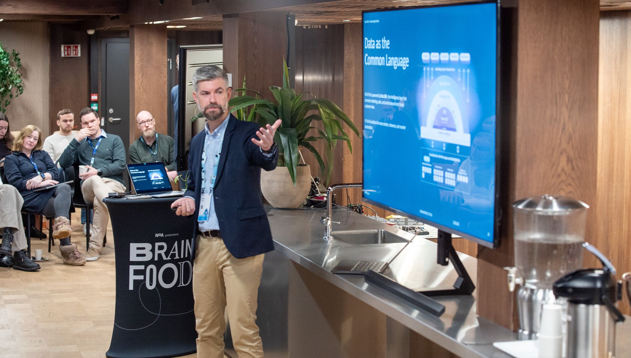 Bearded man in a blazer presenting to a seated audience in a conference room with a large blue display screen.