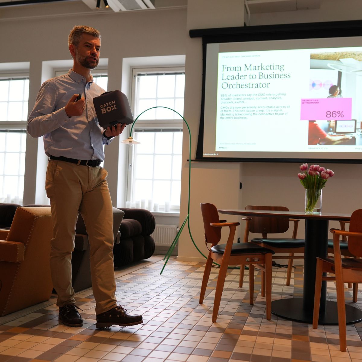 Presenter in a light shirt and khaki pants speaks beside a projection screen showing a slide titled 'From Marketing Leader to Business Orchestrator' while holding a Catch Box microphone in a bright office.